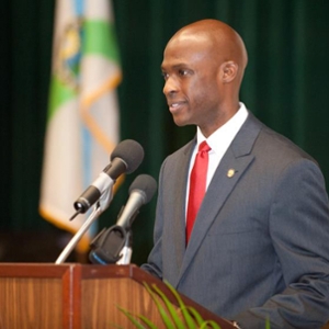 Suited man speaking behind a wooden podium with a flag and green curtain in the background