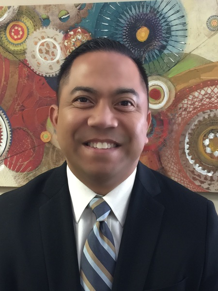 Photograph of a smiling man in a black suit and a striped tie with hanging wall art in the background