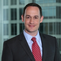 Portrait of a smiling man in a black suit, blue dress shirt and red and white tie with a building in the background