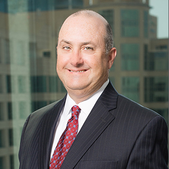 Portrait of a smiling man in a black pinstripe suit standing next to a window with buildings in the background