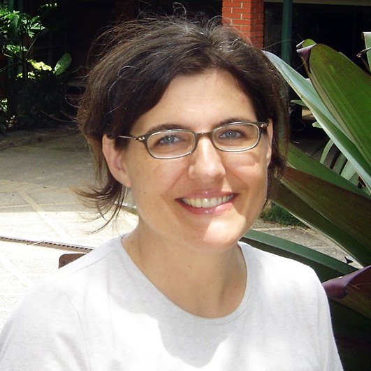 Photograph of a smiling woman wearing glasses and a white shirt outdoors with plants in the background