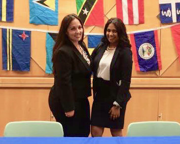 Two smiling women in black suits posing in front of flags hanging on a wall
