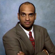 Portrait of a man in a brown suit and red tie with his arms crossed on a gray background