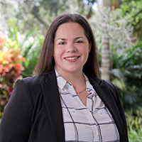 Portrait of a smiling woman in a black suit and white blouse standing in front of green plants