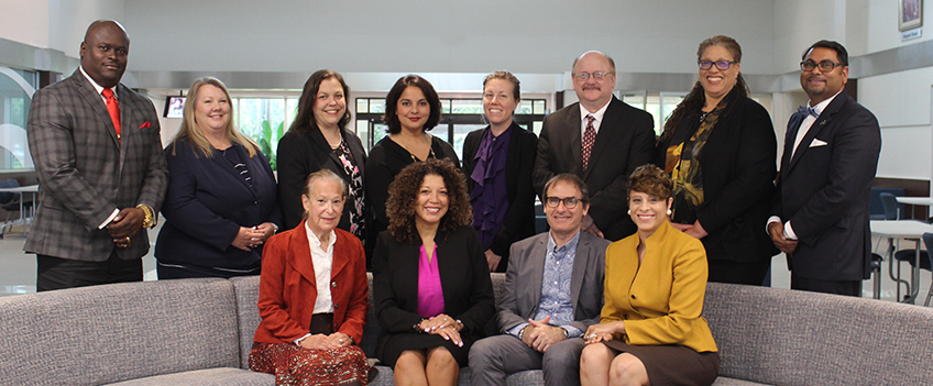 Group picture of smiling adults sitting on a gray couch and posing behind the couch