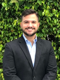 Portrait of a smiling man in a black suit standing in front of green shrubbery